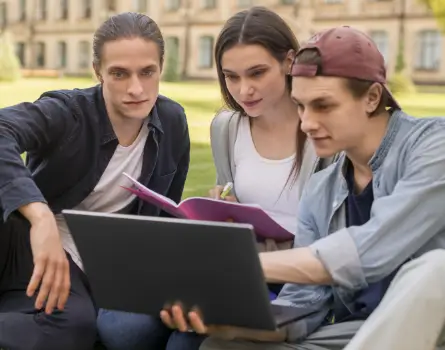 Zwei Studenten und eine Studentin schauen gemeinsam auf ein Laptop. Sie befinden sich auf dem Uni Campus